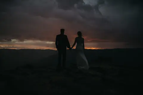 A couple hold hands in silhouette on a ridge beneath a dramatic dusk sky.