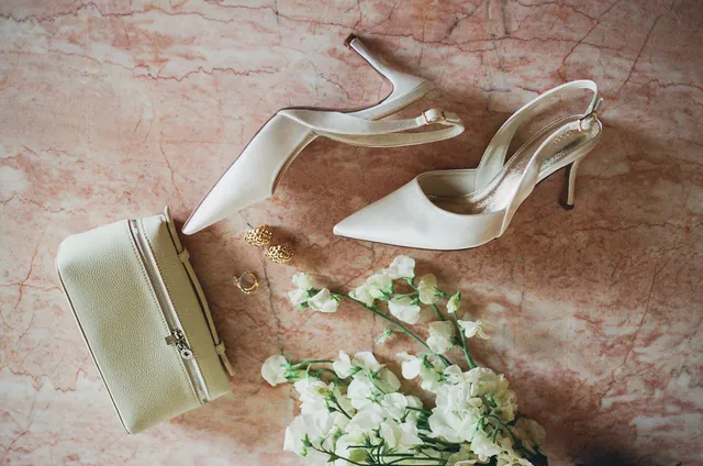 Ivory heels, a pale bag, gold jewellery and white flowers arranged on pink stone.