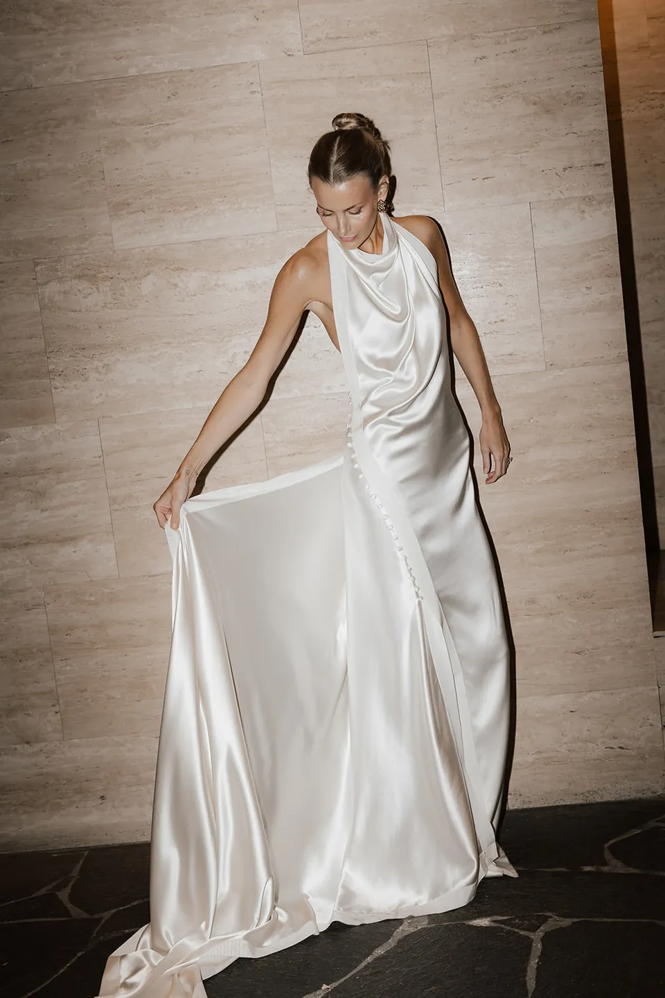 Clare looking down at her flowing ivory dress beside a warm stone wall at The Calile.